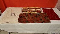 Four Christmas tablecloths folded and laid out side by side on a white surface with red curtain background.
