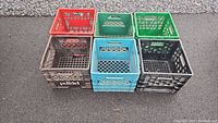 Full view of 6 plastic milk crates of various colors, stacked on ground outside, showing wear and use.