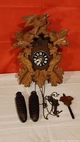Full view of the carved cuckoo clock with two metal pinecone-shaped weights and pendulum leaf on table below it.