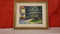 Full view of framed painting showing yellow house with white picket fence and trees in background.