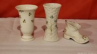 Full view of the three Irish porcelain items: a white shamrock vase, a Belleek basket-pattern vase, and a porcelain shoe, all on display.