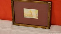 Miniature watercolor painting in gold-colored frame resting against orange wall