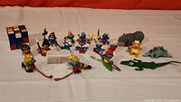 Full wide shot of all figures and toys including Rubik's Cube, Smurf figures in various costumes, and animal figurines arranged on white surface.
