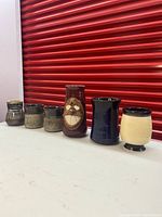 Eight assorted pottery mugs arranged in a row on a table in front of a red background. Variety of sizes, glazes, and styles including a carved face design mug and two matching coffee mugs.