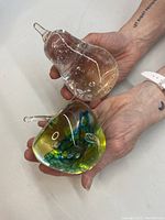 Hands holding clear pear glass art with bubbles and colorful apple glass art with green and yellow swirls.