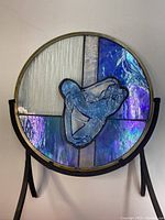 Front view of circular stained glass art piece mounted on black metal stand showing blue and purple glass panels with central abstract 3D glass shape.