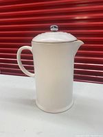 Side view of white ceramic French press with handle and spout, standing on counter with a red background.