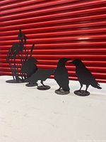 Five black metal bird silhouette sculptures: four crows and one blue heron, arranged on a white surface with a red corrugated backdrop.