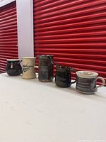 Side view of five pottery mugs in a row showing variety of colors, shapes and glazes