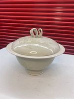 Side-angle photo of off white pottery serving dish with lid featuring a curled handle and textured patch