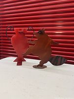 Red metal dancing raccoon sculpture and red metal vase on display against a red corrugated metal background