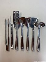 Photo showing seven All-Clad stainless steel kitchen utensils with polished finish including tongs, meat fork, perforated spatulas, solid spatula, and ladle.