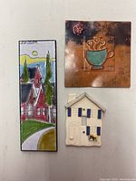 Front view of three pieces of wall art: village scene tile signed Sucsan, pottery house, and copper teacup plate