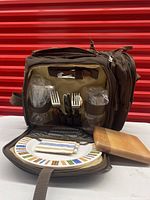 Front view of the open picnic bag showing plates, plastic glasses, utensils, napkins, and wooden cutting board.