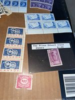 Photo showing multiple Canadian stamps including a block of four blue 5¢ stamps with a bird and a nature scene, a strip of four red 5¢ stamps featuring a maple leaf and hands, one purple $1 Prince Edward Island ferry stamp, and a red $1.44 stamp.