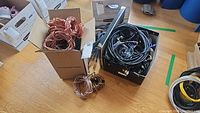 Box containing coiled copper speaker wires and another box with black and other colored cables on a wooden floor.