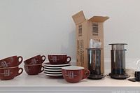 Six brown Cafferty Vita cups and saucers neatly stacked with an Aeropress coffee maker in original box.