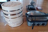 Photo showing three-tier bamboo steamer with metal bands standing next to Cuisinart Griddler with attached power cord on a wooden floor