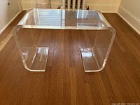 Clear lucite side table on wood floor showing full U-shape design and smooth rounded edges.