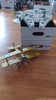 Photo showing the yellow metal biplane model on the floor in front of a box, with the Millennium Falcon model resting on the box.