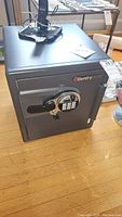 Front view of the SentrySafe with combination keypad, key lock, and handle, placed on wooden floor with additional items on top.