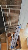 White metal wire shelf leaned against a wall next to a cardboard box containing two roll-up window shades.