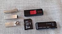 Five storage devices on fabric background: one SanDisk Ultra II 2GB SD card, two SanDisk Cruzer 128GB USB flash drives, two Samsung metallic USB flash drives (128GB and 64GB).