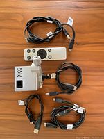 Silver mini projector with ventilation holes and adjustable mounting stand, accompanied by remote control and cables laid out on a wooden surface.