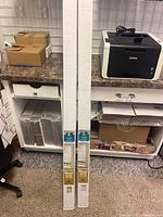 Two boxed Hampton Bay cordless vinyl blinds standing vertically on the floor against cabinets, showing packaging size and general appearance.