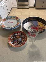 Lot showing six Coca Cola plates stacked, one plate with multicharacter vintage ad design, one metal serving tray with vintage Coca Cola women ads, and one Coca Cola branded glass.