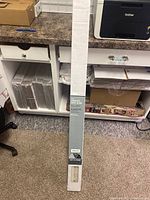 Full block box of the cordless vinyl blind placed vertically against a cabinet and printer, showing packaging and surrounding environment.