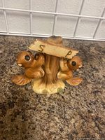 Front view of two squirrels hanging from a tree stump with a wooden sign marked S and P above them on a countertop.