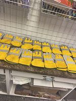 Eighteen pairs of yellow and grey rugged leather gloves arranged in rows showing front sides and some back sides on a counter with a metal grid background.