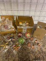 Photo showing multiple Christmas ornaments packed in three boxes and several ornaments laid out. Ornaments are new and wrapped in plastic, colors include gold, red, green, and copper tones with varied shapes like balls and stars.