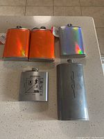 Top view of five metal flasks on table. Three flasks with orange and silver holographic finishes appear new with tags. Two flasks are silver metal with printed and embossed logos, showing used condition.