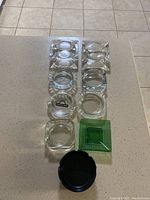 Full layout of 9 ashtrays on a counter, showing all pieces in lot including clear glass, green glass, and black plastic ashtrays.