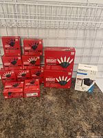 Multiple boxes of red-packaged Christmas lights with 100 bulbs per box, some multi color and some clear, stacked on a floor surface near a wire grid.