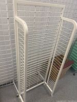 White metal wire rack with grid panels and tubular frame sides, photographed from a side angle showing its height and structure.