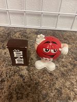 Photo showing both M&M salt and pepper shakers on a countertop near a white grid wall. The red character shaker is raising one arm.