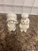 Front view photo of the pair of Mr and Mrs Pillsbury salt and pepper shakers on a kitchen counter with white grid tile background.
