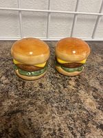 Front view of a pair of ceramic salt and pepper shakers shaped like hamburgers, showing details of buns, lettuce, cheese, and patty.