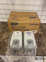 Two Purell instant hand sanitizer dispensers in front of the original packaging box showing branding and model number.