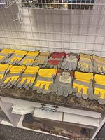 Sixteen pairs of yellow and grey, and red and grey rugged leather gloves displayed on a counter under a metal grid wall.
