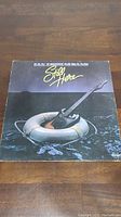 Front cover of the Ian Thomas Band vinyl LP album 'Still Here' showing album artwork with a guitar on a lifebuoy on water.