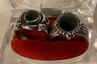 Top view of two men's silver-tone rings with dark colored stones placed side by side on red velvet inside clear case.