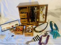 Photo of wooden 4-drawer jewelry box with glass door open showing hanging necklaces and some drawers partially open, various pieces of necklaces and bracelets laid out around box.