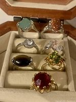 Seven ladies rings with various colored stones displayed in a cream padded jewelry box with wooden frame, showing different sizes and shapes of stones in both silver and gold-tone settings.