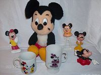 Seven vintage Mickey and Minnie Mouse collectibles arranged on a white cloth backdrop, including plush toy, figurines, baby rattle, and cups.