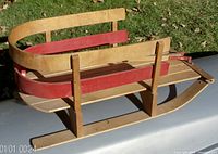 Side and front angle view of the vintage child's wooden winter sleigh showing curved wooden rails, red accent paint, seat base, and handle.