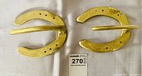 Top view showing front side of two gold-colored horseshoe shaped coat hangers on white fabric background.